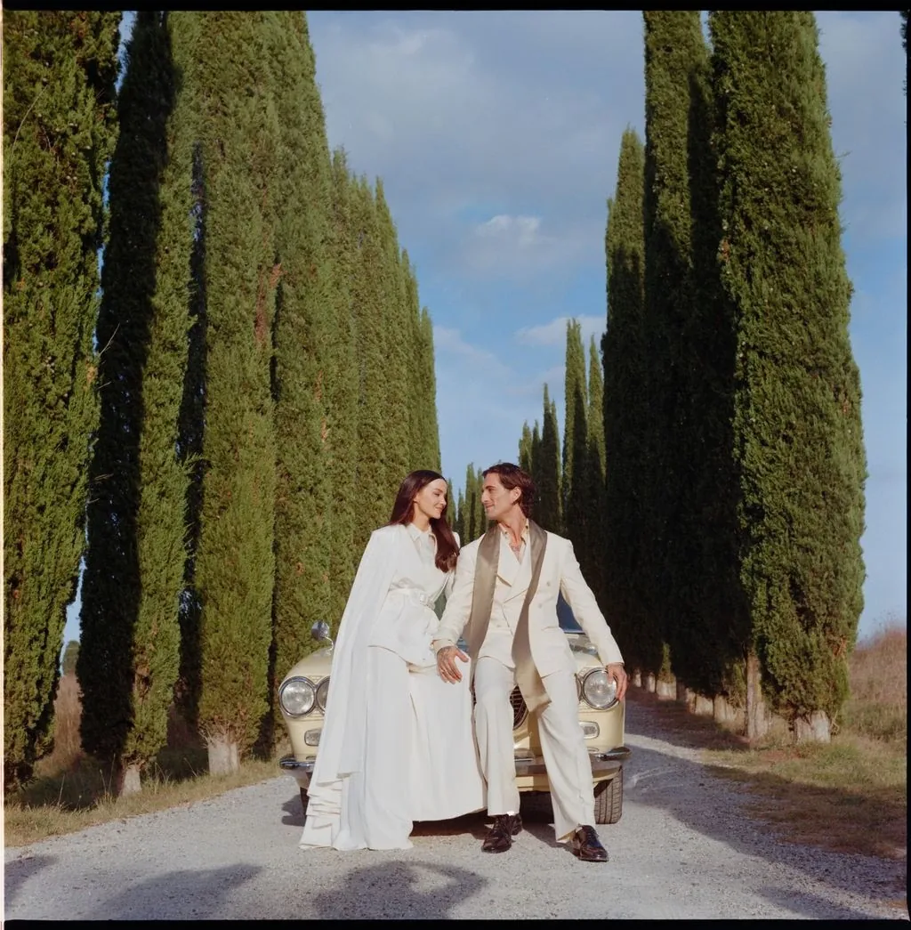 Dove Cameron and Damiano David in white tailoring beside a vintage car in Tuscany
